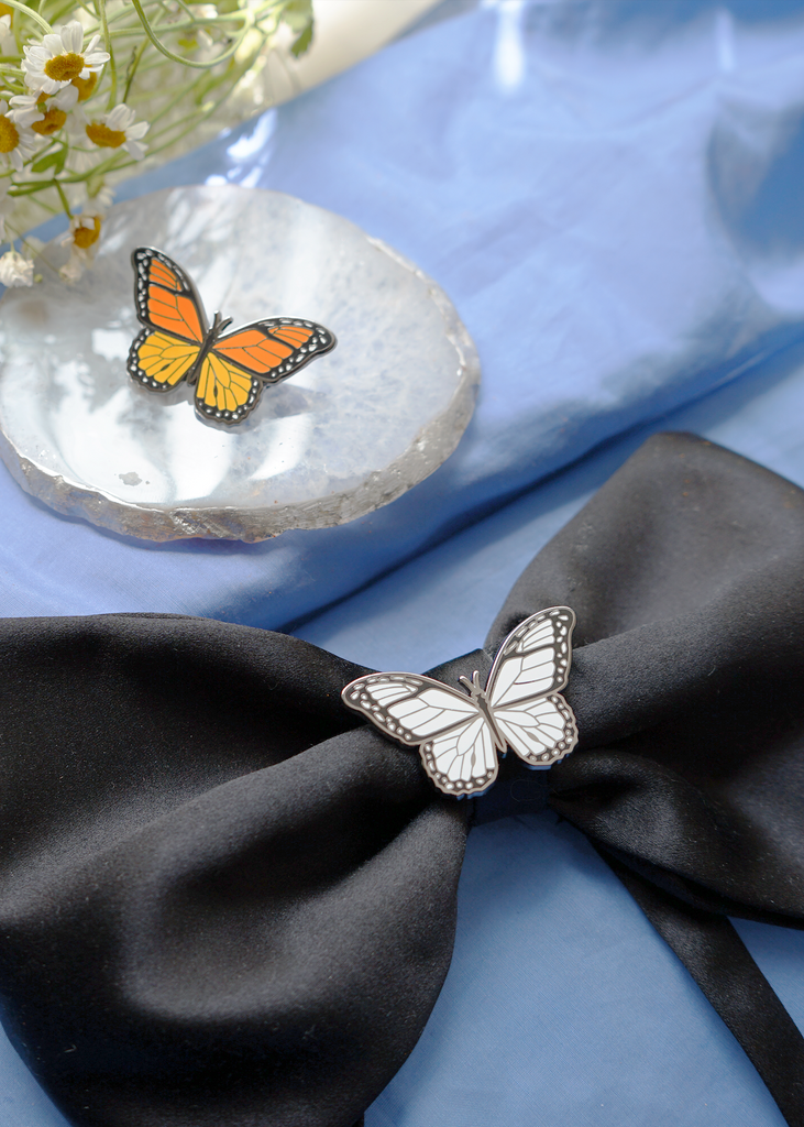 White Monarch pin on Black satin bow next to orange monarch enamel pin placed on agate stone (pins sold separately)