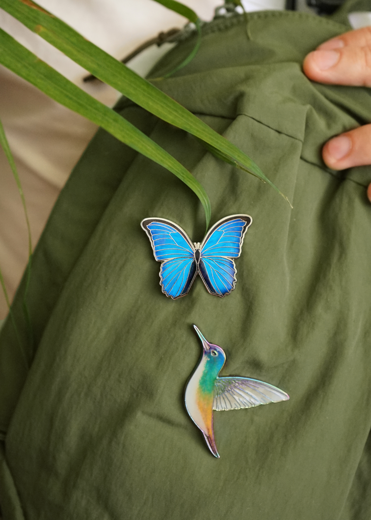 Godart's blue morpho pin on green crossbody next to buff bellied hummingbird pin